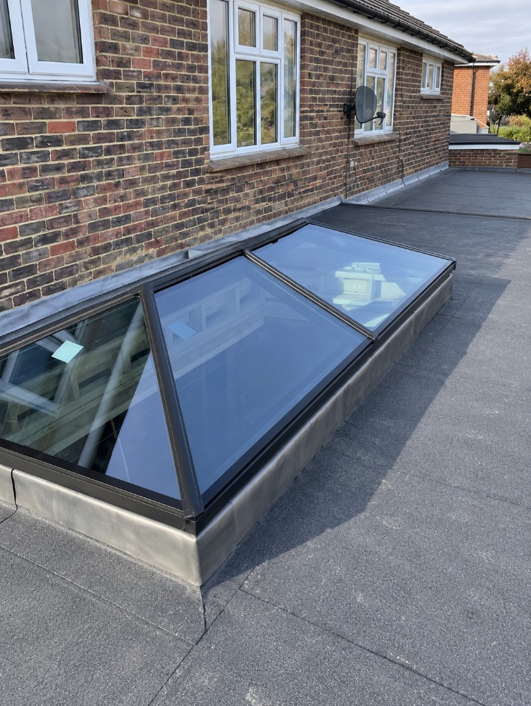 Black-framed roof lantern on a grey flat roof beside a brick house